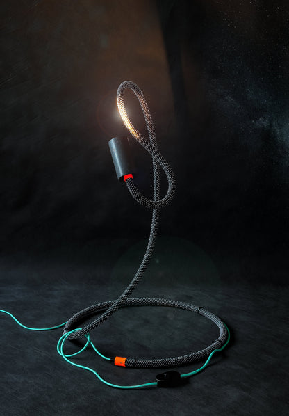 Leuchte NAGA in flippiger Ausführung. Mit schwarz-weiß schraffierter Ummantelung und türkis metalic Stoffkabel. Die Akzente der Leuchte sind Neon-Orange.