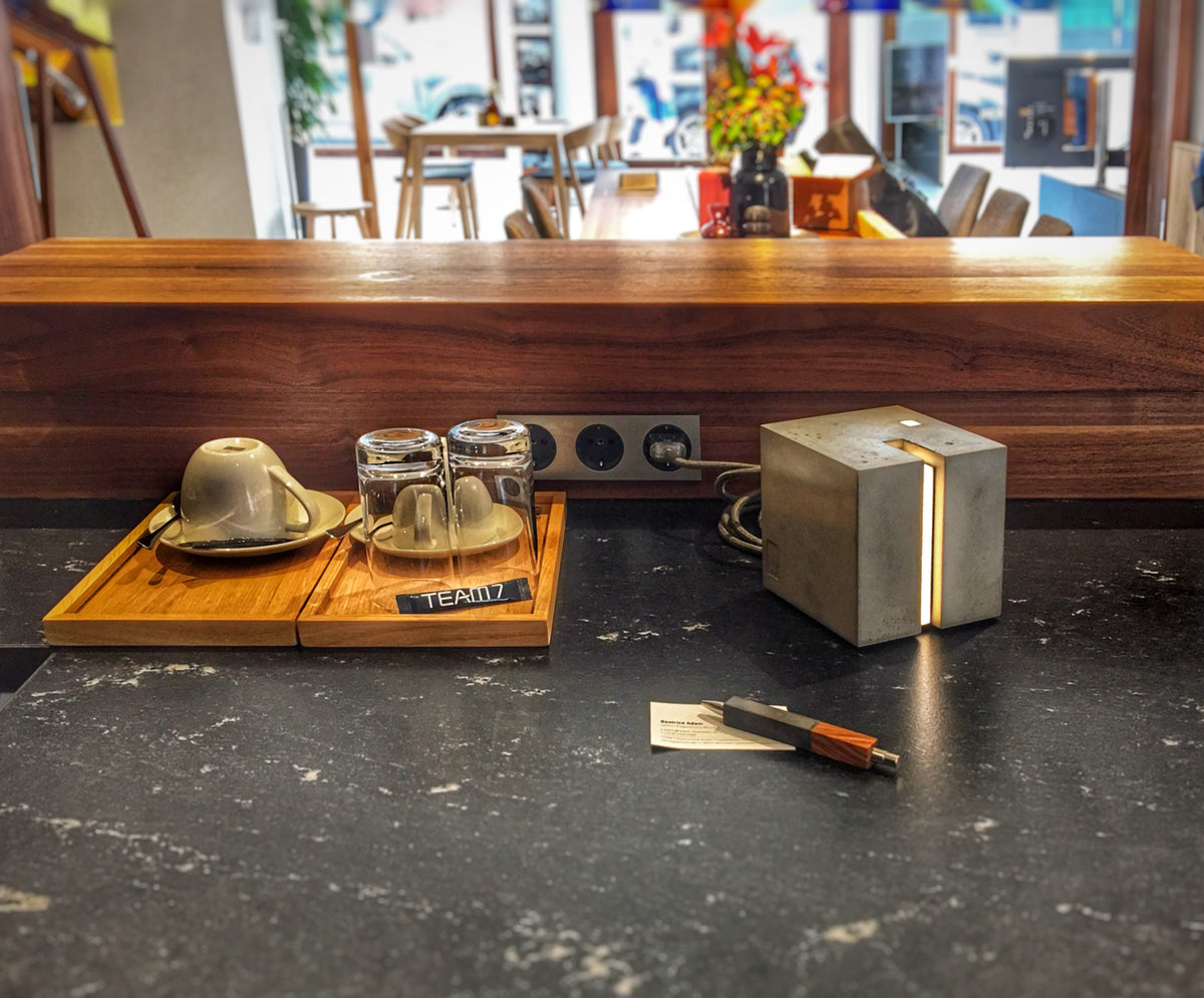 Eine Beton Leuchte in form eines Würfels auf einem Tisch. Darum herum ein Kugelschreiber und Tassen und Gläser. In dem Hintergrund ist unscharf der Rest des RAumes zu sehen